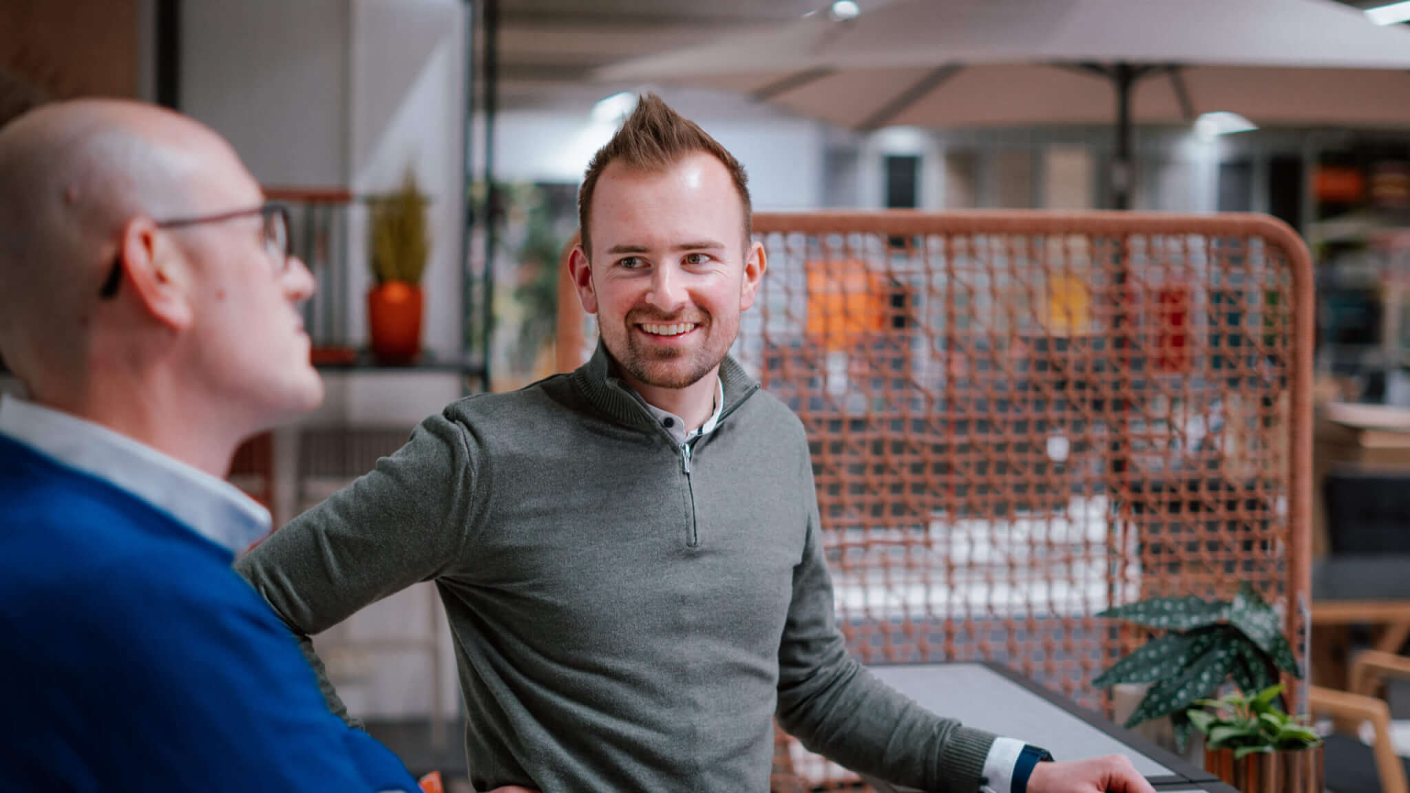 Ein Mann im Fokus steht mit einem Kollegen zusammen an einem schmalen Tisch.